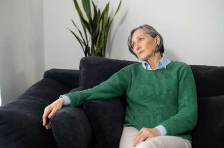 Senior mature lady is sitting on the couch in the living room and looking in the window, feeling tired fatigued, having no energy and no motivation to get up, having a bad day conceptの写真素材