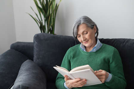 Portrait of a charming retired senior mature lady wearing green sweater jumper and reading an interesting book with curiosity, sitting on the sofa or couch in the living room at homeの写真素材