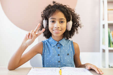Video chat with a pretty curly African schoolgirl wearing a headphones, she looks into camera, shows victory gesture and smiles. A pretty biracial girl talking via video call with classmatesの写真素材