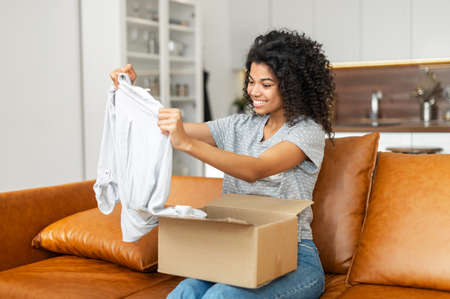 Satisfied smiling young African American woman sitting on the sofa opening parcel carton box,...
