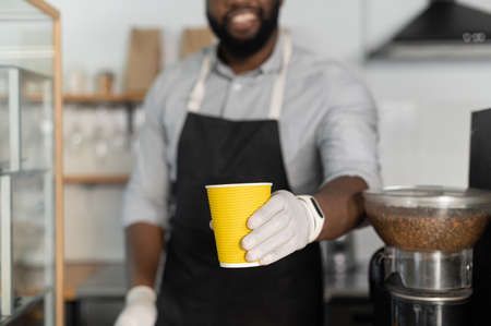 Cropped picture a multiracial barista wearing gloves and apron serves customer, giving a paper cup, holds out a coffee to goの写真素材