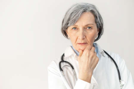 Professional look of a serious senior mature female doctor with a stethoscope in a medical coat, posing isolated over gray background, placed her hand on a chin, looking at the cameraの写真素材