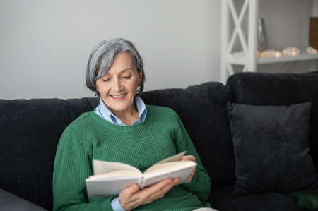 Happy grandmother senior mature woman in a green sweater, sitting on the couch and reading a book,...