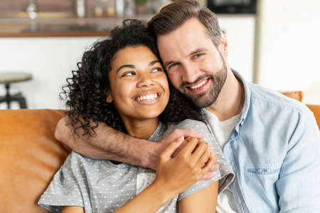Hilarious multiracial couple in love sits in embraces in cozy living room at home. A close-up portrait of smiling caucasian guy and charming african girl are hugging. Love and affection conceptの写真素材