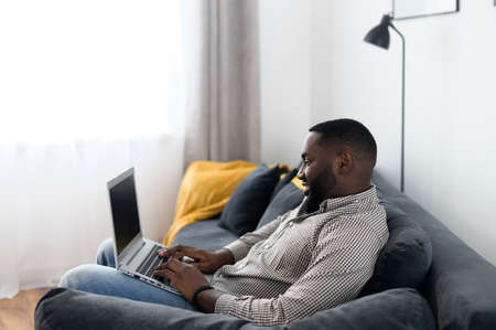 Young attractive bearded African American guy placed the laptop on knees while relaxing on the sofa, typing email, messaging friend, freelancer working from home on a project, chatting, dating onlineの写真素材