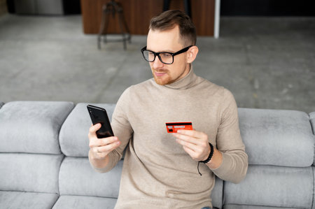 Cheerful young man makes purchase online. A guy wearing casual shirt holds a mobile phone and a...