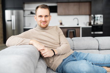 Portrait of handsome young man having rest in modern living room at home, sitting casually on sofa, looks at the camera, feeling relaxed. Lifestyle and leisureの写真素材