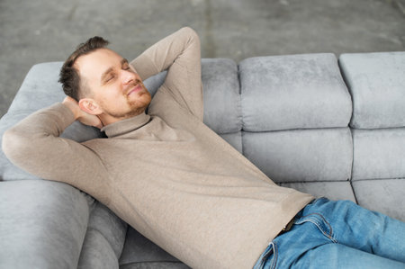 Handsome smiling guy take a nap lying on the comfortable sofa with eyes closed and hands behind head, attractive bearded man rests, daydreaming, restingの写真素材