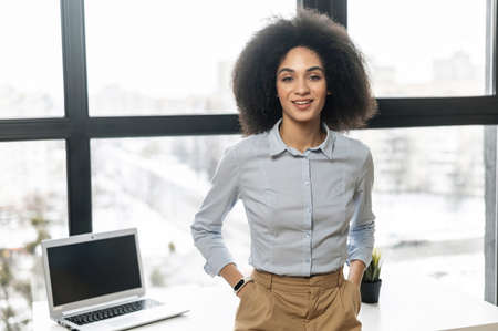 Successful confident young African American female entrepreneur businesswoman with Afro hairstyle in casual clothes standing with hands in pockets against window with panoramic view, looking at cameraの写真素材