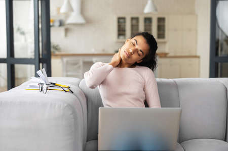 African American female freelancer or student with curly hair sitting on the couch in modern living room, feeling tired from constant work on laptop, removed her glasses, stretching neck with handの写真素材