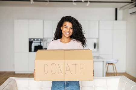 Young pretty cheerful African American woman smiling standing in the living room and holding carton box for donation and donate is written by hand, mixed-race girl looking at camera, charity conceptの写真素材