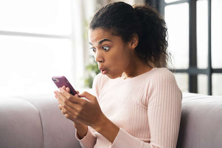 Surprised African American teenage girl sitting on the couch, looking at mobile phone, checking social media messages or browsing the internet and cannot believe her eyes, unbelievable news conceptの写真素材