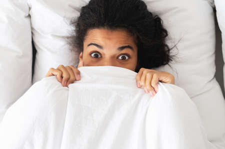Surprised, shocked biracial woman with wide open brown eyes, dark hair and bronze skin while lying in the bed, covering half of face with white blanket, top view, afraid of nightmares, after awakeningの写真素材