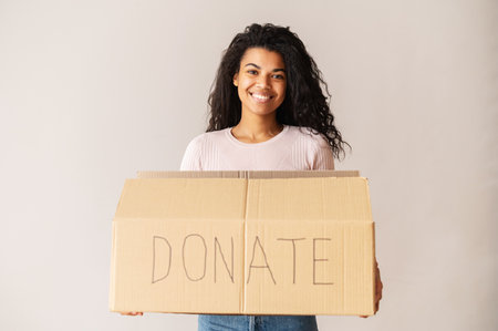 Kind smiling African American female volunteer with curly dark hair holding a cardboard box, donating her things to charity, for orphans and homeless, for reuse or recycle of clothes or toysの写真素材