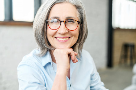 Headshot portrait of a charming beaming elegant mature woman with her hand on a chin, wearing glasses and looking at the camera. Close-up of a middle-aged gray-haired female office worker or a teacherの写真素材