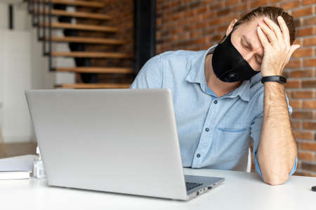Disappointed businessman with hair pulled up, in mask, holding the head, looking at the computer screen, crisis in the company, working online, checking the balance on the bank account, use antisepticの写真素材