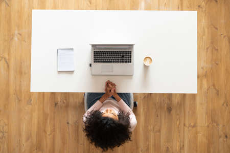 Overhead view of African American woman or female office worker with curly hair sitting at the modern white table desk with well-organized space, in front of a laptop, taking notes, ready to workの写真素材