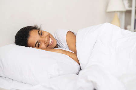 Positive young joyful relaxed African American girl covered with soft blanket lying down and staying at home, resting head on the pillow and looking at the camera, enjoying a day off or weekend in bedの写真素材