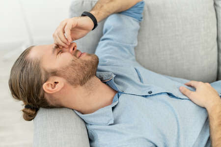 Attractive man with light brown hair pulled back, beard, lying on the grey cozy couch, with pleasure smile, keeping arm on the face, remembering this fantastic day, got long-awaited job, after dateの写真素材