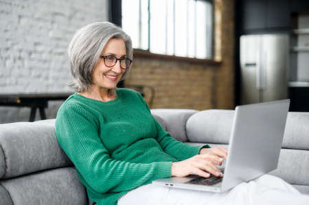 Cheerful senior woman sitting on the couch in cozy living room at home, using computer laptop for spending leisure time, remote working. Middle-aged lady with answering emails, writes an articleの写真素材