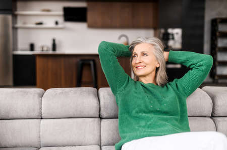 The winsome senior lady is resting on the comfortable couch at home, charming elderly woman in casual wear put her hands behind her head and daydreaming in relaxed atmosphere, wellbeing conceptの写真素材