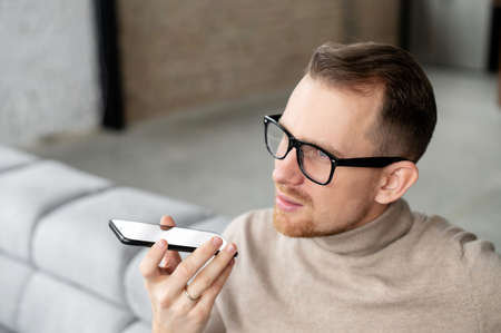 Close up of handsome married bearded hipster man entrepreneur businessman in glasses holding mobile phone sitting on the couch in a living room, talking, recording a voice message or asking directionの写真素材