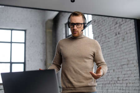 Portrait of young businessman in a headset looking on the laptop, hipster customer service representative standing in the modern office, actively thinking and concentrating on the customers problemの写真素材