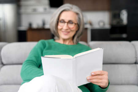 Thoughtful happy mid-aged senior woman in glasses and green jumper sitting on the couch, resting and...