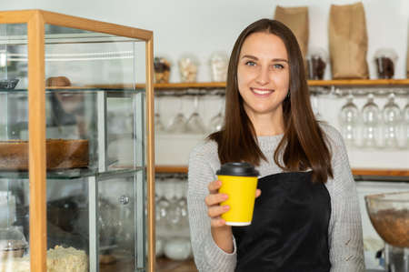 Friendly and smiling waitress in apron holds out paper cup of take away coffee into the camera....