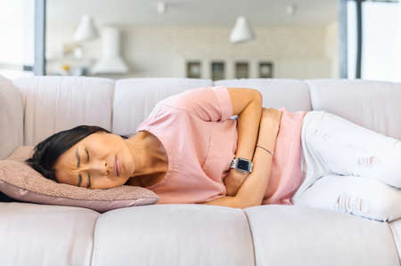 A young Asian woman lying on the couch and suffers from month menstruation period pain, stomach...