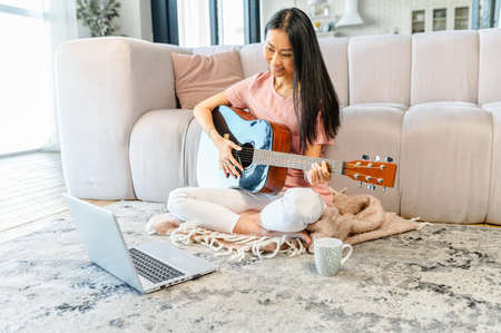 Hobby and e-study concept. A young Asian woman is watching on the laptop video tutorial how to play guitar, sits with an acoustic guitar on the floor in cozy living room and repeat exercisesの写真素材