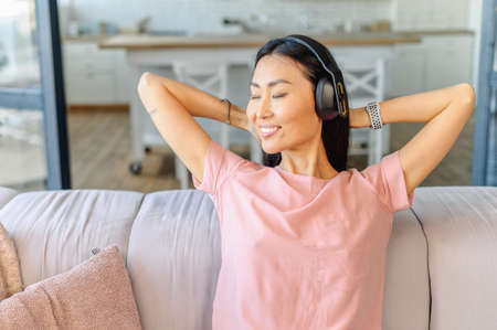 A satisfied and cheerful young Asian woman wearing headphones sits on the couch, rests at home...