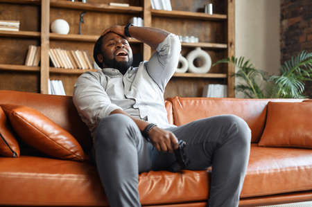 Frustrated African-American guy lost in video game, a young man sitting on the couch with a...