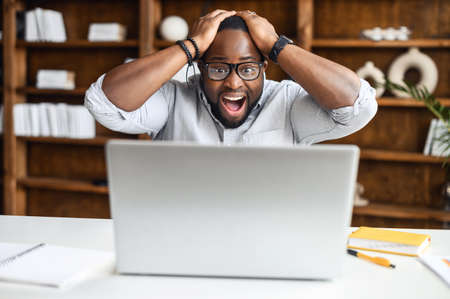Shocked man is staring on the laptop screen, holding head and yelling happily, surprised...