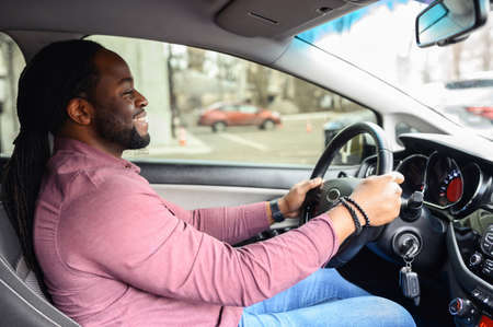 A happy African-American man with the dreadlocks is driving a car, side view from a passenger seat...