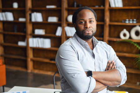 Portrait of a serious young confident African American male entrepreneur standing with arms folded in the library or and looking at the camera, intelligent black IT guy posing with arms crossedの写真素材