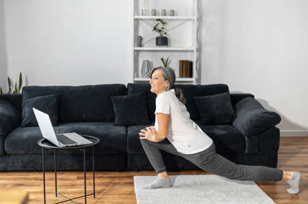 Cheerful and smiling mature woman watching fitness and yoga classes for senior on the laptop, an elderly female in sportswear stretching legs at home, leads a healthy and active lifestyleの写真素材
