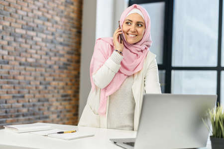 Business muslim woman in hijab at office, standing at the desk with laptop, talking by mobile phone,...