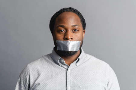 Serious African-American man with with a sealed mouth with tape looks at the camera sadness, speechless, speechless, racism concept, isolated on greyの写真素材