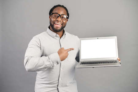 Cheerful African-American businessman wearing smart casual shirt and eyeglasses demonstrates a laptop with a blank screen, a guy points finger at empty computer monitor, mockup, isolated on greyの写真素材