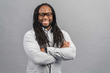 Confident cheerful African-American with long dreadlocks wearing smart casual shirt stands with arms...