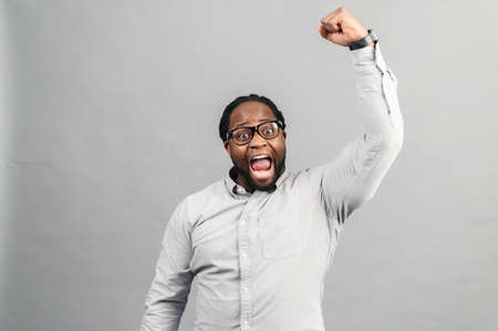Excited African-American guy wearing smart casual shirt celebrating victory, raising clenched fist up, screams yes happily, black guy yelling ecstatic, triumph concept, studio shot isolated on greyの写真素材