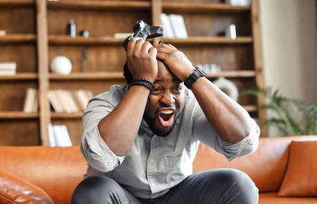 Front view portrait of young African American guy sitting on the couch in the living room, holding head in hands and joystick, screaming because lost video game match, male addicted gamer feeling madの写真素材