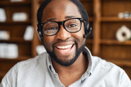 Smiling young African-American salesman in glasses and headset looking at the camera and laughing, positive black man on video call working in the customer service department and talking onlineの写真素材