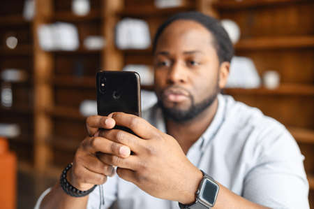 Suspicious man taking photo of criminal act or serious African American hodling mobile phone and typing text message, reading bad news, young upset black guy looking at the cellphone screenの写真素材