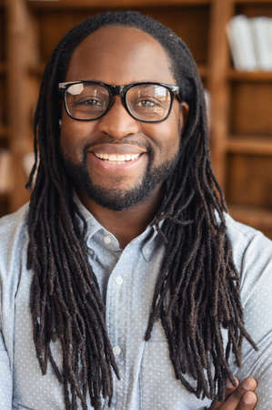 Vertical portrait of smiling young handsome African American man with dreadlocks and glasses standing with arms folded and looking at the camera, optimistic black man office manager is ready to workの写真素材