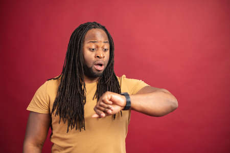 Amazed surprised young African American man with dreadlocks isolated on red wall forgot about business meeting, checking time from watch on his wrist, going to be late again, time for changes conceptの写真素材