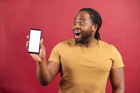 Surprised by the possibilities of new mobile app African-American man staring at smartphone with empty screen, astonished dark skinned guy demonstrates phone with blank display, isolated on redの写真素材