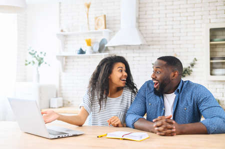 Excited multi ethnic couple in love does not believe their eyes, sits at the kitchen table in front to a laptop, looks to each other with shock, received mortgage approval by mail, win in lotteryの写真素材