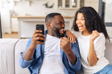 Excited and overjoyed African-American young couple sits on the couch at home and holds smartphone, a man and woman show victory gesture, celebrating good news or win, received money on accountの写真素材